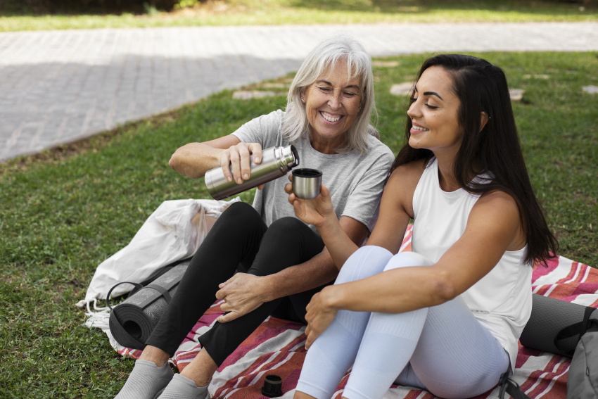 Mlajša in starejša ženska skupaj sedita v parku po vadbi joge in si delita čaj – simbolni prikaz solidarnosti, na kateri temelji pokojninski sistem.