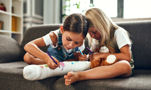 Simbolna fotografija za nezgodno zavarovanje otrok in mladine - dve deklici rišeta na mavec deklice s poškodovano nogo