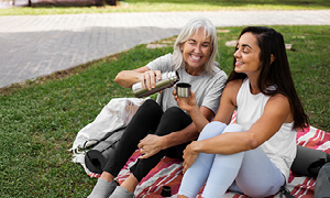 Mlajša in starejša ženska skupaj sedita v parku po vadbi joge in si delita čaj – simbolni prikaz solidarnosti, na kateri temelji pokojninski sistem.