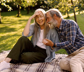 Dva starejša občana sedita na piknik odeji v senčnem mestnem parku; travnik in drevesa v poletnem popoldnevu.