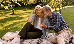 Dva starejša občana sedita na piknik odeji v senčnem mestnem parku; travnik in drevesa v poletnem popoldnevu.