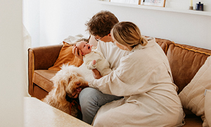 Mlad par sedi na kavču v dnevni sobi in pestuje majhnega otroka, ob njiju je pes z dolgo dlako; prizor prikazuje domačo družinsko bližino.